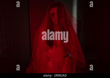 Scena horror di una sposa posseduta. Donna con abito bianco zombie fantasma che tiene in mano bouquet di rose essiccate in una stanza buia con luce rossa. Halloween spaventoso Foto Stock