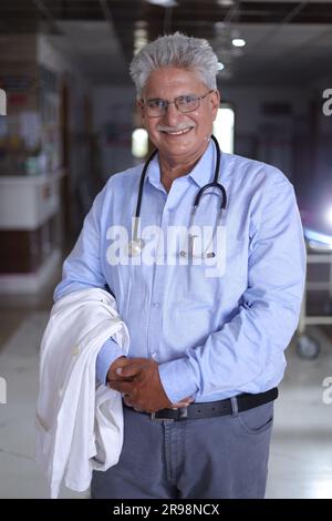 Felice indiano senior medico ritratto in piedi in corridoio di ospedale vestito con scrub e camici da laboratorio, con stetoscopi. Foto Stock