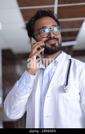 Ritratto medico professionale in corridoio di ospedale vestito in scrub e camici da laboratorio, con stetoscopi. Indiano decente che parla su un cellulare. Foto Stock