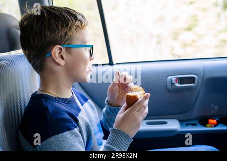 Un felice ragazzo caucasico in età scolare cavalca sul sedile posteriore di una macchina e mangia un panino. Uno scolaro con gli occhiali. Il bambino indossa le cinture di sicurezza, AN Foto Stock