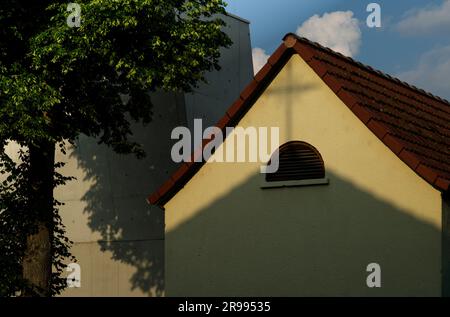 L'ombra di una croce di chiesa gettata su un timpano di giallo chiaro con una finestra semicircolare sotto un edificio in cemento e un albero verde Foto Stock