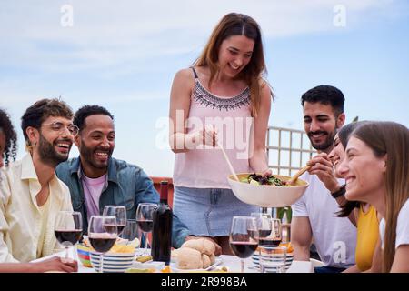 Donna caucasica riunita con i suoi amici, serve insalata ai suoi ospiti. Gruppo di adulti in terrazza. Foto Stock