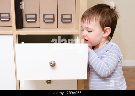 Il bambino ha aperto il cassetto del mobiletto. Il bambino gioca con il guardaroba nel salotto di casa. Bambino di un anno e nove mesi Foto Stock