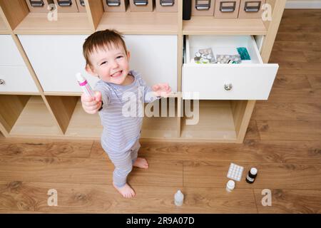 Il bambino ha aperto il cassetto con pillole e unguento. Bambino che tiene la crema in piedi nel salotto di casa. Bambino di un anno e nove mesi Foto Stock