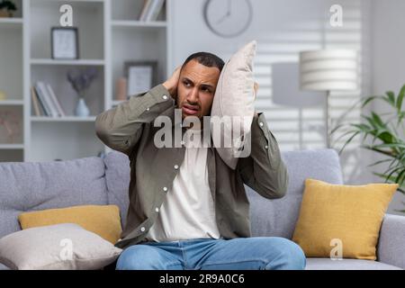 Un giovane afro-americano è seduto sul divano a casa e si copre le orecchie con cuscini. Arrabbiato soffre di rumore eccessivo da parte dei vicini, riparazioni. Soffre di attacchi di panico, voci. Foto Stock