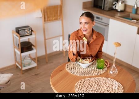 Delizie nitide. Signora felice che si gode la sua insalata caesar preparata in casa, seduto al tavolo in cucina, spazio libero, vista dall'alto Foto Stock