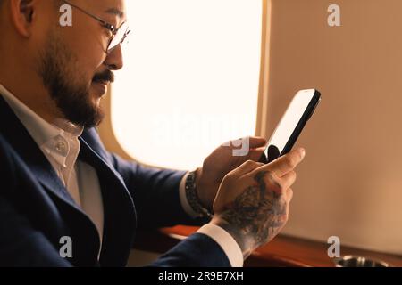 Elegante giovane uomo d'affari asiatico, con occhiali da vista e tuta, seduto all'interno in jet privato di prima classe mentre lavora con uno smartphone moderno Foto Stock