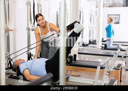 Allenatrice sportiva e giovane che controlla i movimenti degli uomini anziani che si allenano con l'attrezzatura del letto Pilates Reformer in palestra Foto Stock