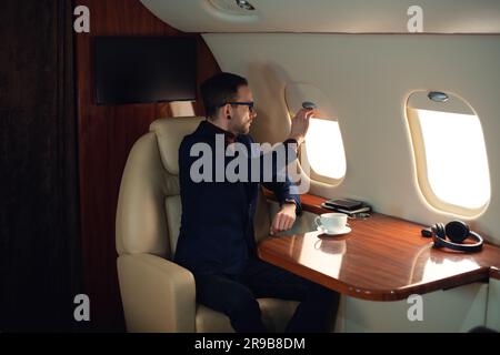 Elegante uomo d'affari adulto con occhiali da vista abbassa il paralume della finestra prima di decollare con un jet privato da lavoro, uomo d'affari sicuro di sé con una giacca blu Foto Stock