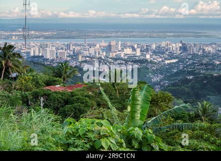 Prima del tramonto la luce del sole bagna gli edifici più alti della capitale di Cebu, guardando attraverso il canale del mare fino all'Isola di Maktan, sopra le palme e il lussureggiante fogliame Foto Stock