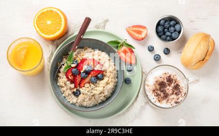 Porridge di farinata d'avena con fragole, mandorle e banana in ciotola. Colazione salutare con cappuccino e succo d'arancia su fondo bianco. Vista dall'alto. Foto Stock