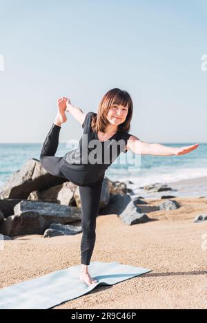 Donna matura in abbigliamento sportivo in equilibrio sulla gamba a Natarajasana. Si trova sul tappetino mentre fa yoga sulla costa sabbiosa vicino al mare nelle giornate di sole e guarda la macchina fotografica Foto Stock