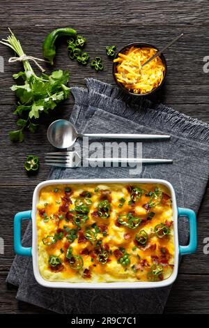 Schiacciate di patate cariche con formaggio cheddar, formaggio spalmabile, pancetta e panna in piatto da forno su un tavolo di legno scuro, vista verticale Foto Stock