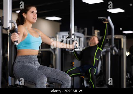 Giovane ragazza concentrata che conduce uno stile di vita sano e attivo facendo allenamento di forza in palestra, eseguendo esercizio di farfalla per i muscoli toracici in pec deck m Foto Stock