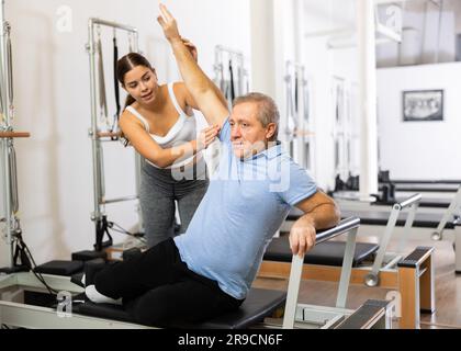 Allenatrice sportiva e giovane che controlla i movimenti degli uomini anziani che si allenano con l'attrezzatura del letto Pilates Reformer in palestra Foto Stock