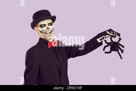 Un uomo allegro in costume di Halloween su sfondo lilla leggero posa con la tarantola giocattolo in mano. Foto Stock