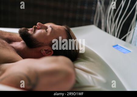 Vista dall'alto del bel giovane sdraiato rilassandosi sul letto con massaggio elettrico tenendo le mani dietro la testa, godendo di un trattamento spa nel lussuoso centro benessere Foto Stock