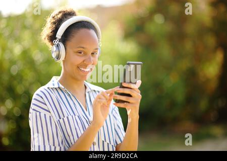 Donna nera felice che indossa le cuffie e regge lo smartphone che posa guardando la fotocamera mentre ascolta la musica in un parco Foto Stock
