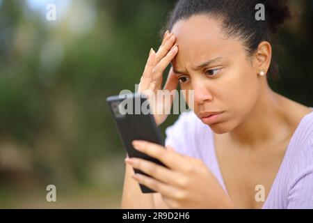 Preoccupata donna nera che controlla cattive notizie sul cellulare in un parco Foto Stock