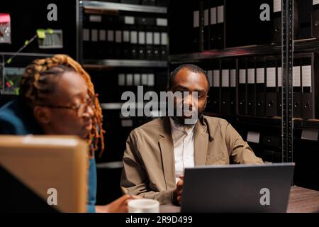 Poliziotta afroamericana che aiuta il detective con l'analisi delle prove. Investigatori che analizzano gli indizi di casi criminali su un computer portatile mentre lavorano straordinari nell'archivio della polizia di notte Foto Stock