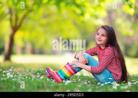 Ragazza carina toddler in stivali di gomma da pioggia giocare all'esterno nel parco verde con fioritura campo di fiori margherite, ritratto di bambino Foto Stock