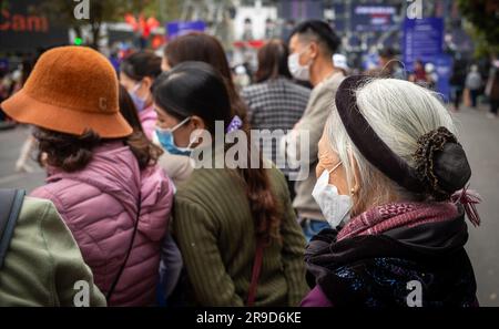 Una donna anziana che indossa abiti tradizionali e una maschera facciale sta accanto ai giovani ad Hanoi, in Vietnam. Foto Stock