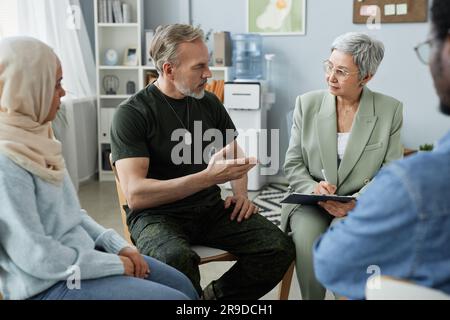 Uomo maturo barbuto in abbigliamento casuale che descrive il suo problema allo psicologo mentre sedeva tra gli altri pazienti durante la comunicazione durante la sessione Foto Stock