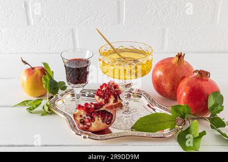 Una ciotola di miele, un bicchiere di vino rosso, melograni maturi e mele su un vassoio d'argento contro una parete di mattoni bianchi. Il concetto di Rosh Hashanah Foto Stock
