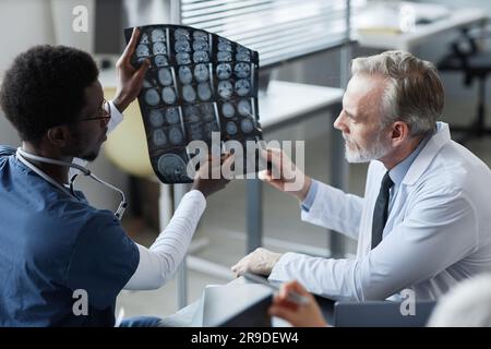 Giovane assistente afro-americano che mostra e spiega i risultati della risonanza magnetica del cervello del paziente al radiologo maschio maturo in studio medico Foto Stock