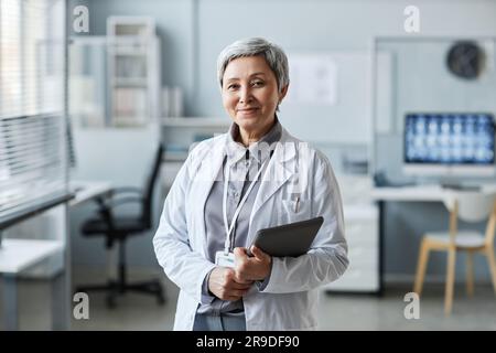Felice maturo medico con tablet in piedi di fronte alla macchina fotografica nello studio medico e guardando contro i posti di lavoro dei colleghi Foto Stock