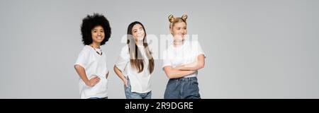 Ragazze adolescenti gioiose e multietniche con magliette bianche che posa e guarda la fotocamera mentre si trovano l'una accanto all'altra isolate su un grigio e vivace t Foto Stock