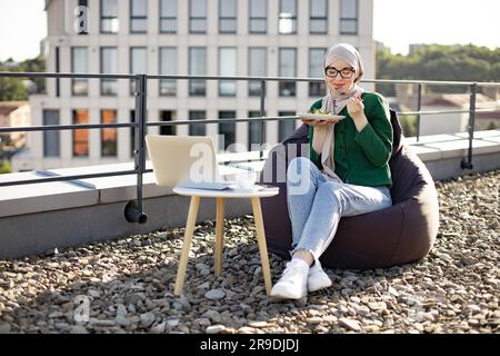 Giovane donna che indossa hijab e bicchieri che mangia cibo sano mentre è seduta in una sedia beanbag sul tetto dell'edificio della città. Una donna araba positiva che fa una pausa pranzo durante un lavoro remoto su un computer portatile all'aperto. Foto Stock