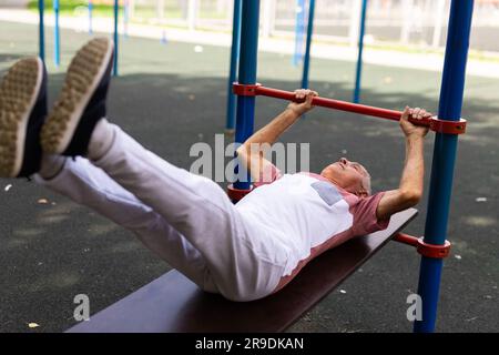 L'uomo anziano pompa i muscoli addominali e brucia le calorie. Atleta anziano attivo che fa esercizio fisico su campi sportivi, allenandosi in palestra all'aperto Foto Stock