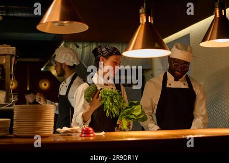 Giovane donna sorridente in uniforme che porta broccoli freschi e verdure a foglia verde allo chef tritando ingredienti per insalata o altro piatto Foto Stock