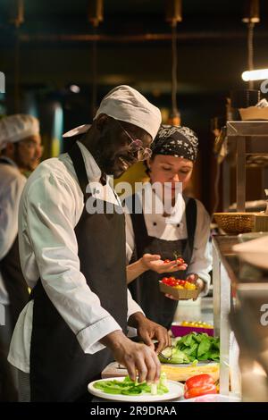 Felice giovane chef afroamericano in uniforme, mescolando ingredienti freschi su una piccola tavola rotonda prima di metterli in padella Foto Stock