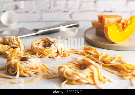 Nidi di tagliatelle di zucca, alcune fette di zucca in un piatto, conchiglie d'uovo e un coltello su fondo di marmo. Foto Stock