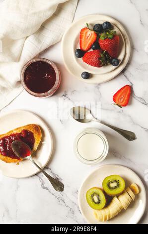 Yogurt semplice, frutta fresca, marmellata di fragole in un vaso e su una fetta di pane tostato con un tovagliolo bianco su fondo marmorizzato. Vista dall'alto. Foto Stock