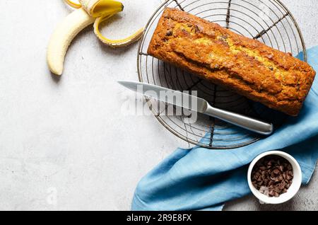 Un pane alla banana con gocce di cioccolato, una banana, gocce di cioccolato in una ciotola e un tovagliolo blu, su sfondo grigio. Foto Stock