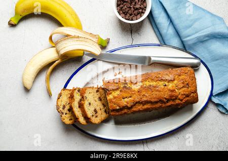 Un pane alla banana affettato con gocce di cioccolato, banane, gocce di cioccolato in una ciotola e un tovagliolo blu, su sfondo grigio. Foto Stock