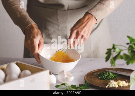 Primo piano delle mani femminili che sbattono le uova in un recipiente. Prezzemolo e aglio su una tavola di legno. Il processo di preparazione della milanica argentina. Foto Stock