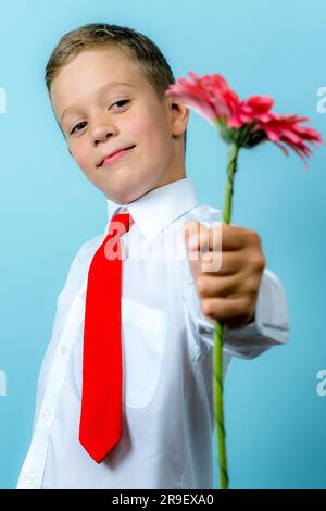 un primo elementare felice con una camicia bianca tiene un fiore nelle sue mani e te lo consegna. Un bel ragazzo caucasico va a scuola con un mazzo di fiori. Foto Stock