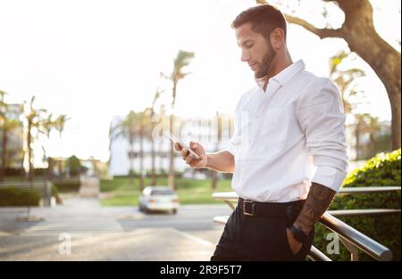 Giovane uomo d'affari che indossa abiti formali in piedi all'aperto e utilizza il cellulare, chiacchierando con clienti o partner commerciali Foto Stock