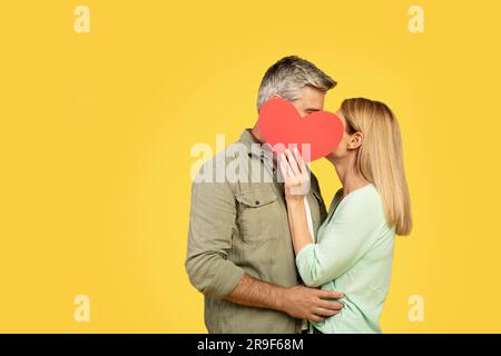 Innamorati di mezza età che si nascondono dietro il cuore di carta rossa e si baciano, coprendo i loro volti, sfondo giallo, spazio libero Foto Stock