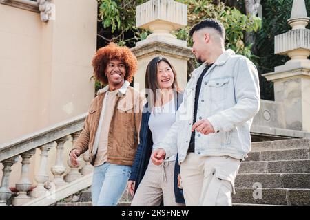 Gruppo di studenti adolescenti che camminano giù per le scale, parlano e sorridono nel campus universitario. Tre giovani amici multirazziali che visitano e si divertono insieme. Gente allegra che si fa beffe del fine settimana. Foto di alta qualità Foto Stock