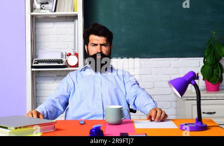 Insegnante di sesso maschile con la barba seria in classe. Apprendimento e istruzione. Giornata degli insegnanti. Insegnante professionista o professore universitario seduto al tavolo con Foto Stock