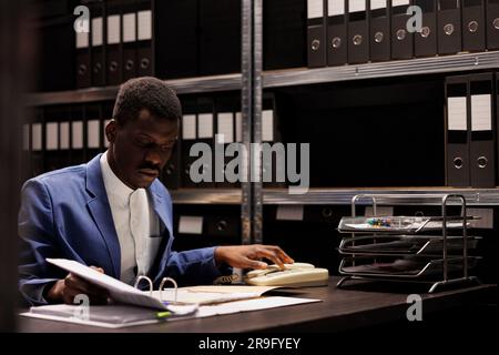 Ispettore afroamericano che risponde al telefono, discutendo le prove investigative con un agente di polizia a distanza che usa la linea fissa in sala aralveare. Detective privato che lavora a tarda notte al caso penale Foto Stock