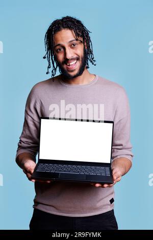 Allegro uomo arabo in piedi con un notebook che mostra uno schermo vuoto per un ritratto simulato su una pagina Web. Persona in possesso di un computer portatile con display bianco per la pubblicità e guardare la fotocamera Foto Stock