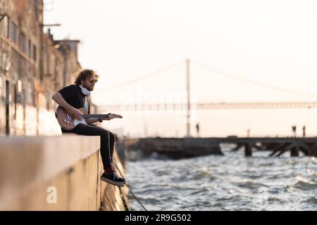 Un uomo hipster siede sul terrapieno con le gambe appese all'acqua e suona la chitarra. Scatto medio Foto Stock