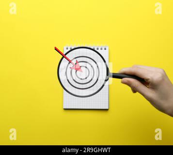 Donna che tiene una lente di ingrandimento sopra un notebook su sfondo giallo, vista dall'alto. Immagine del bersaglio e della freccia Foto Stock