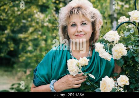 una donna di 59 anni in giardino. Una donna matura gode dei fiori di una rosa coltivata con le proprie mani nel suo giardino. Foto Stock